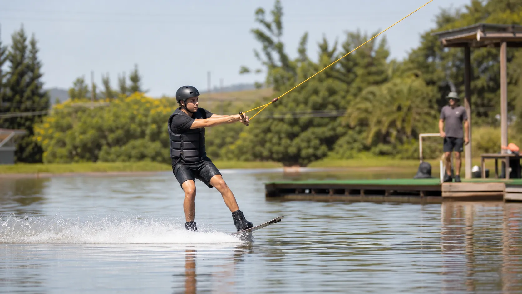 Pessoa iniciante andando de wakeboard em um sistema de cable park com instrutor ao fundo
